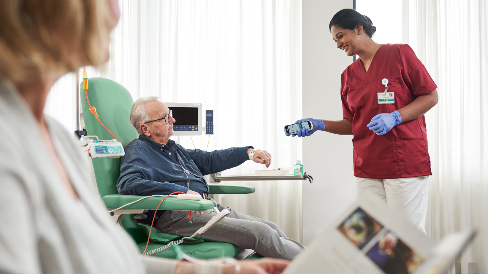 Onkologipatient med briller siddende på stolen og sygeplejerske med handsker, der scanner patientidentifikation på armbånd for sikkerhed og dokumentation 