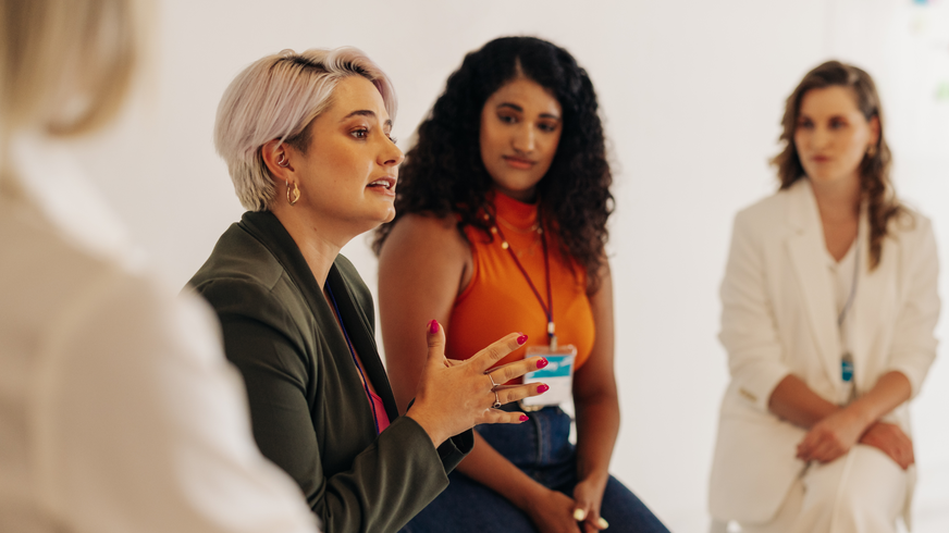 Fire kvinder sidder side om side i Women’s Mentoring Program. Målet er at øge synligheden af kvinder i organisationen og hjælpe dem med deres personlige vækst. En anden kvinde observerer den første, en gråhåret kvinde i grøn jakke, mens hun taler og giver et håndsignal.  