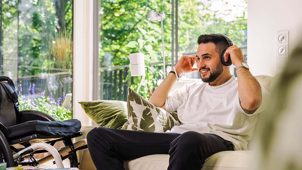 Til højre sidder en mand på sofaen med høretelefoner på, han holder hænderne på høretelefonerne. Til venstre er der en kørestol