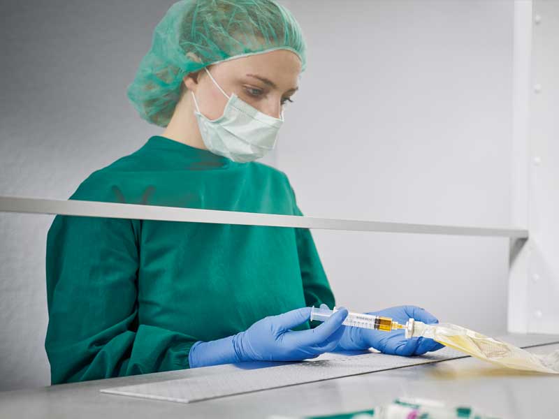 En sygeplejerske iført steril uniform med ansigtsmaske forbereder parenteral ernæring i et sterilt miljø og injicerer en væske i en ernæringspose. Forskellige medicinske artikler er synlige i forgrunden.