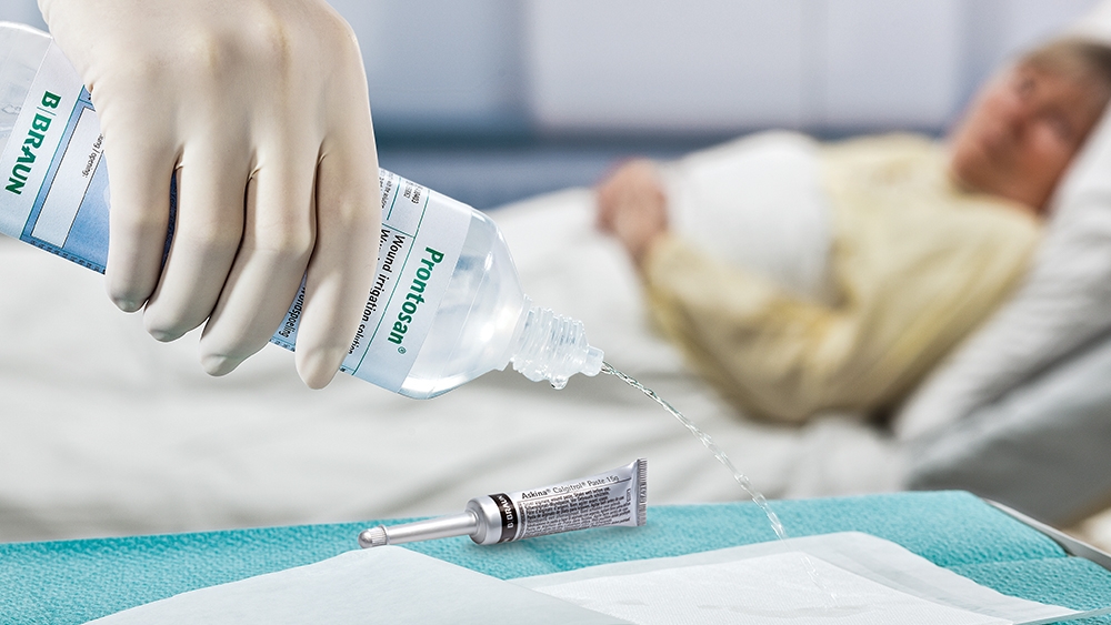 I forgrunden hælder en hånd med medicinsk handske Prontosan Irrigation Solution på en pude, der ligger på bordet ved siden af et Askina Calgitrol-rør. I baggrunden ligger en kvindelig patient i en hospitalsseng.
