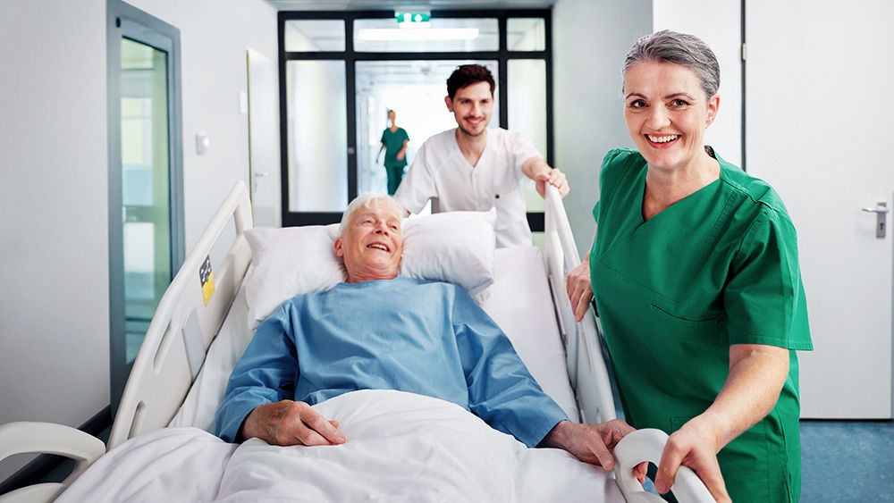 Patient, der ligger i en hospitalsseng og transporteres gennem en korridor af sundhedspersonale, med et andet personalemedlem, der går ved siden af