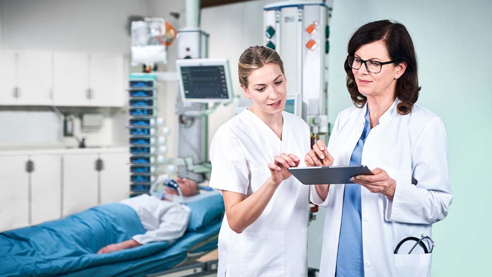 En læge og en sygeplejerske diskuterer doseringen af B. Braun smart infusion, som indtastes direkte fra en tablet. Patienten på intensivafdelingen kan ses i baggrunden, forbundet til et tårn fuldt af smarte infusionsenheder.