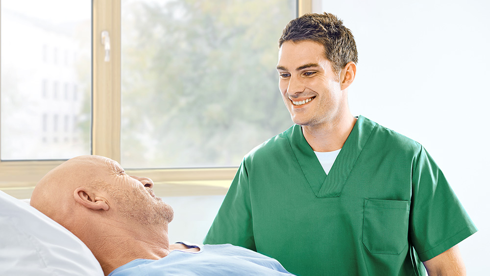 Mandlig sygeplejerske smiler til en patient, der ligger i sengen, hvilket repræsenterer "Mere tid til patientpleje". 