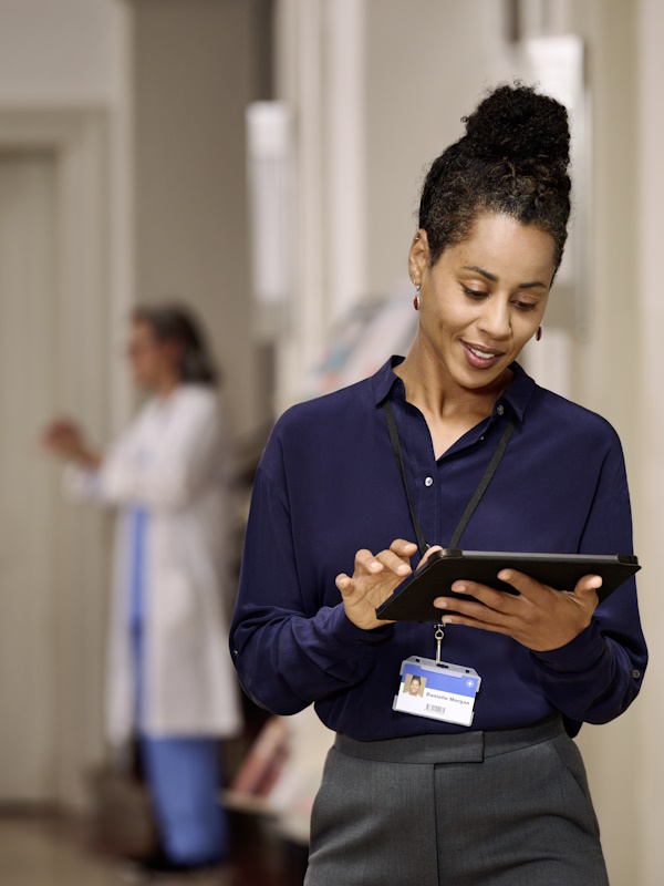 En kvinde i forgrunden, klædt i en mørkeblå skjorte og med et identifikationsbadge på en snor, står i en entré og holder og kigger på en tablet. I baggrunden ses en anden person iført medicinsk tøj.