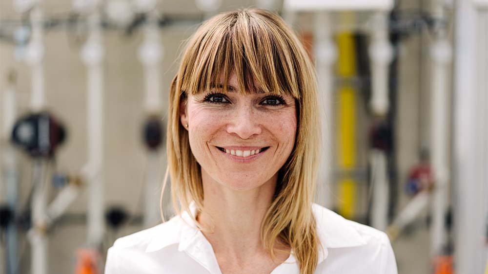 Nærbillede af en kvinde, der kigger ind i kameraet, smiler og har en hvid skjorte på i lyse omgivelser. 